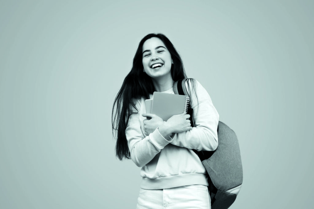 Excited young female student with long dark hair in casual clothes smiling brightly while standing against pink background with backpack and stack of notebooks after successful exam